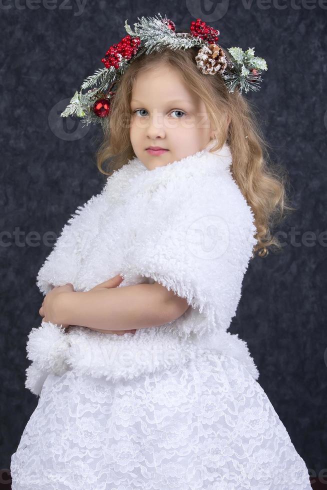 Beautiful little girl in a white fur coat with a Christmas wreath on her head on a gray background. Christmas child. photo