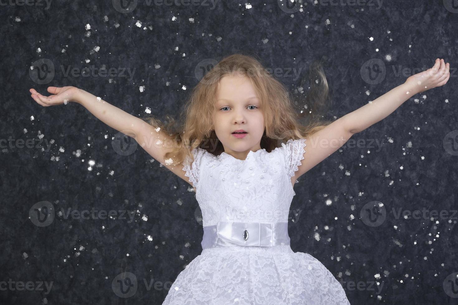 Beautiful little girl in a white dress with raised arms under flying snowflakes. photo