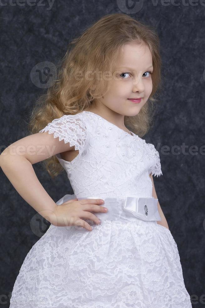 linda pequeño rubia niña en un hermosa blanco vestir en un oscuro antecedentes. seis año antiguo hermosa niña foto