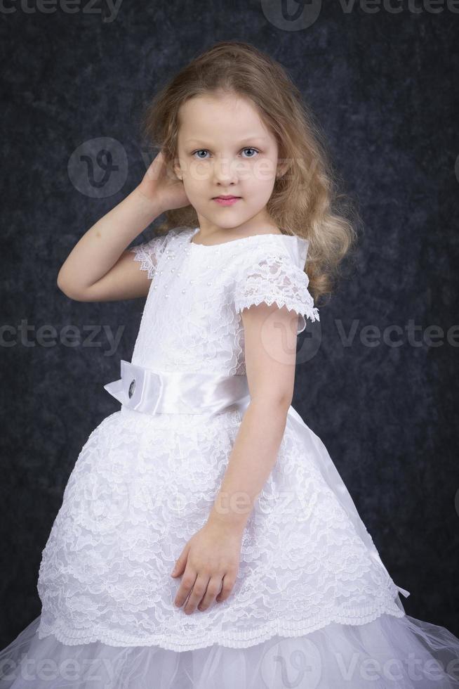 linda pequeño rubia niña en un hermosa blanco vestir en un oscuro antecedentes. seis año antiguo hermosa niña foto