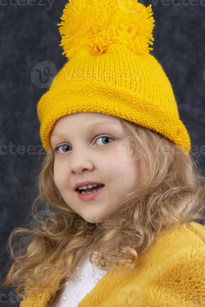 A beautiful little blonde in a knitted yellow hat looks at the camera and smiles. Happy beautiful child about five years old. photo