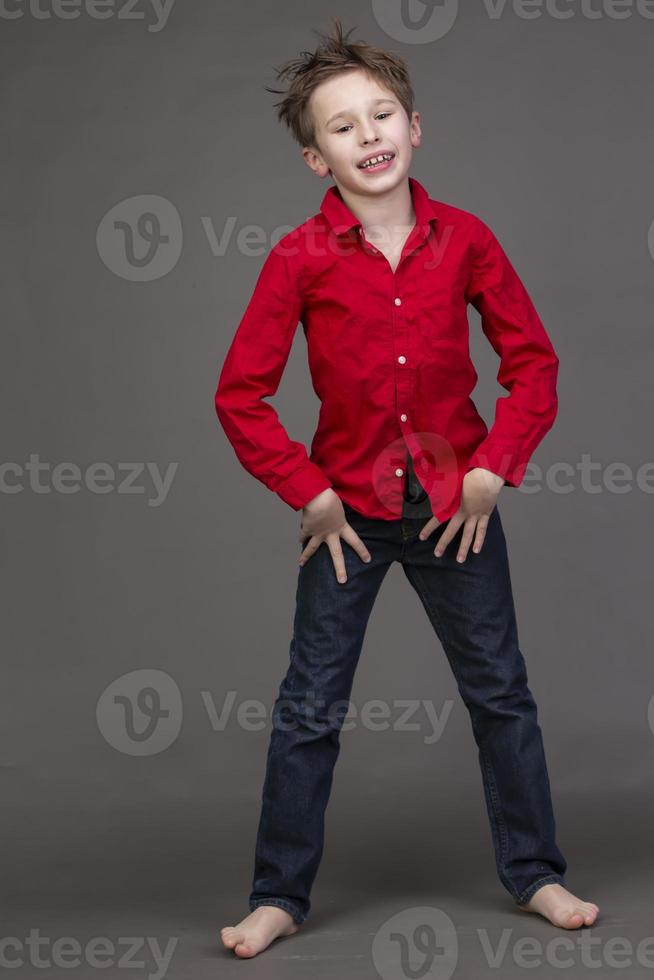 Handsome boy in a red shirt and jeans on a gray background. A child in the modeling business is posing. photo