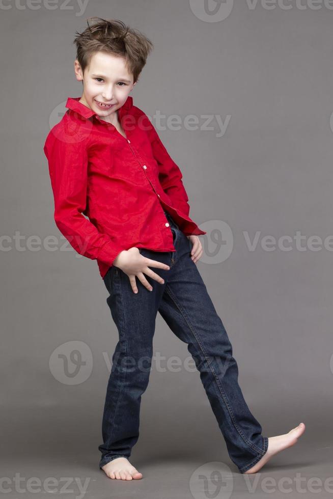 Handsome boy in a red shirt and jeans on a gray background. A child in the modeling business is posing. photo