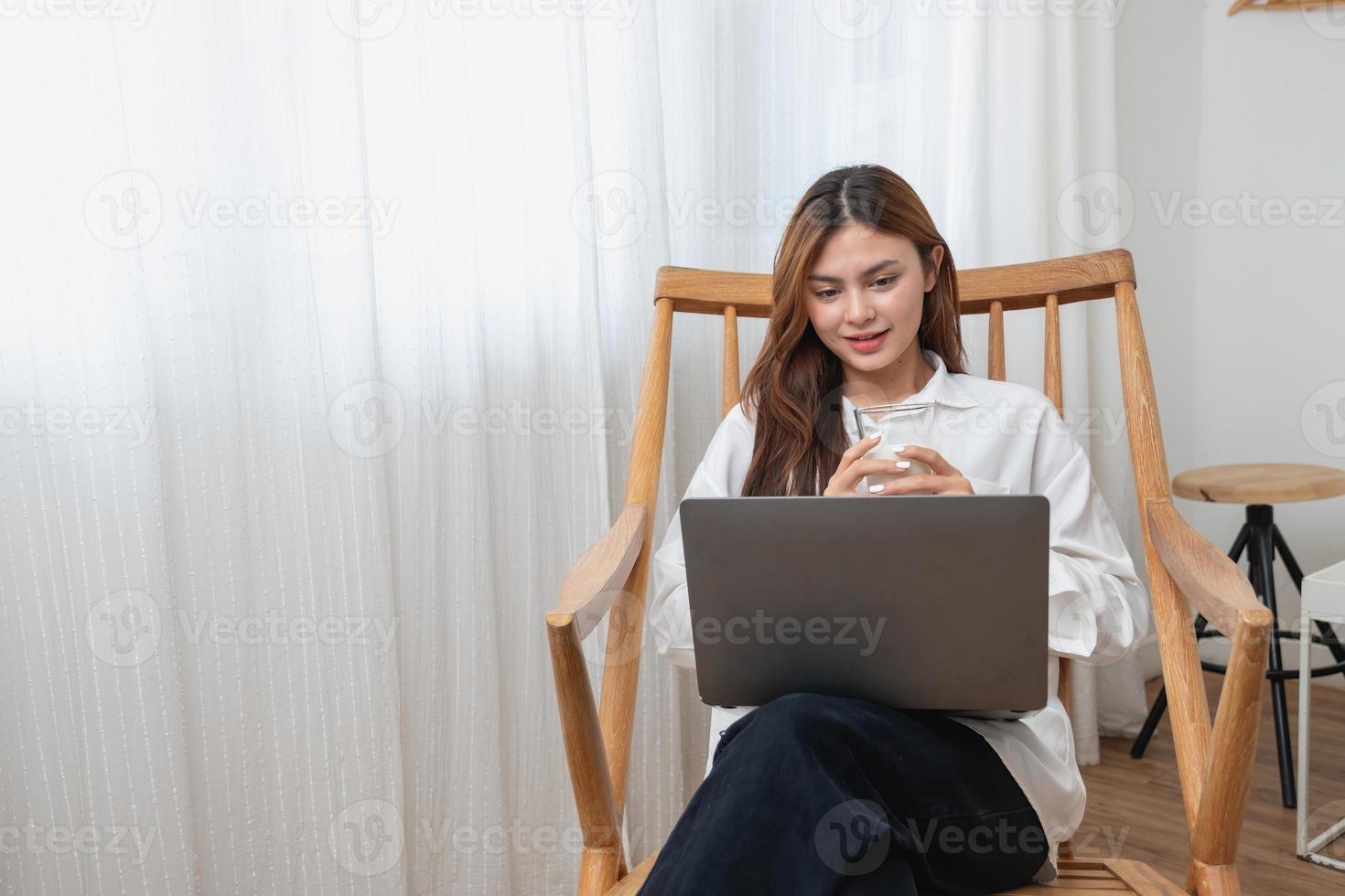 retrato de un hermosa joven mujer en un blanco camisa participación un vaso de Leche y un ordenador portátil a tipo su propio historia y sentado en un de madera silla dentro el casa, el concepto de descanso foto