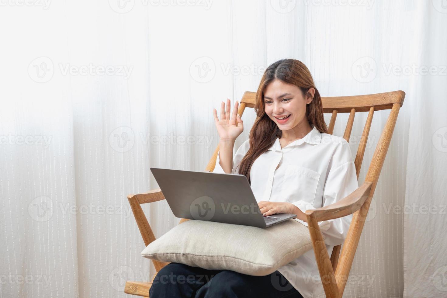 retrato de un hermosa joven mujer vistiendo un blanco camisa participación un ordenador portátil a tipo su propio historia y sentado en un de madera silla dentro el casa, el concepto de descanso y relajación foto