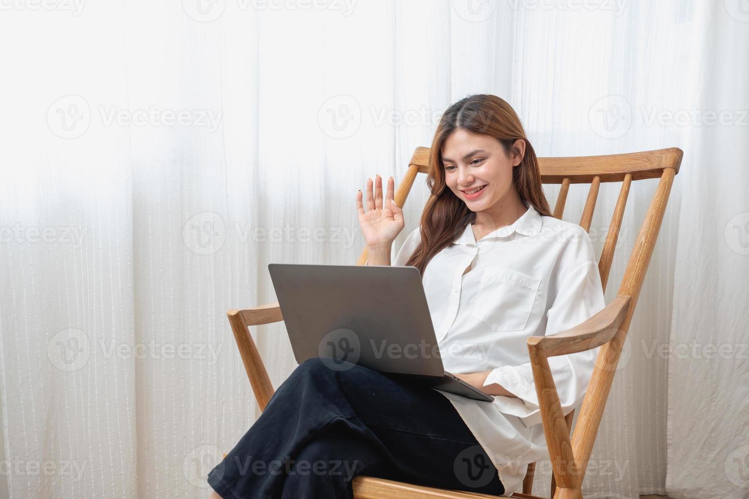 retrato de un hermosa joven mujer vistiendo un blanco camisa participación un ordenador portátil a tipo su propio historia y sentado en un de madera silla dentro el casa, el concepto de descanso y relajación foto