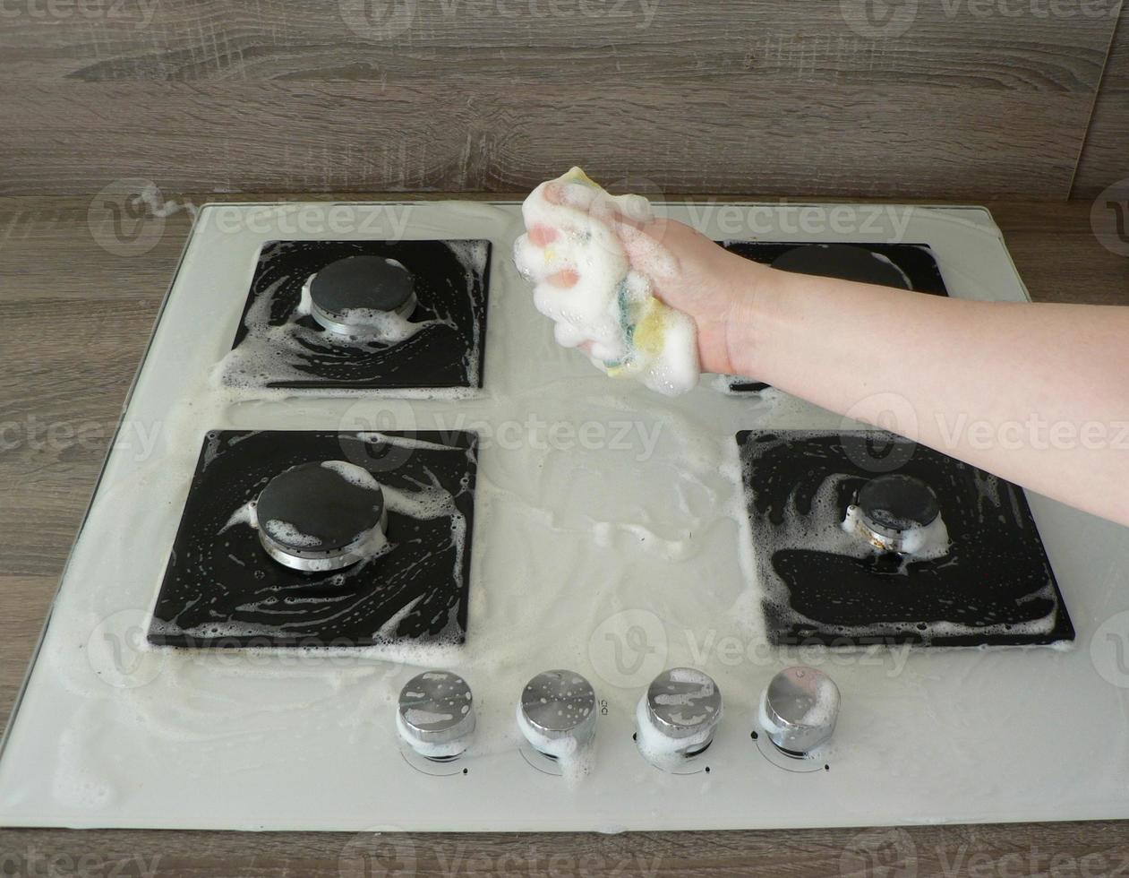 Cleaning dirty gas stove, foam from household chemicals. Hands hold a washing sponge in the foam. White glass hob surface. The concept of household chores, house cleaning. photo