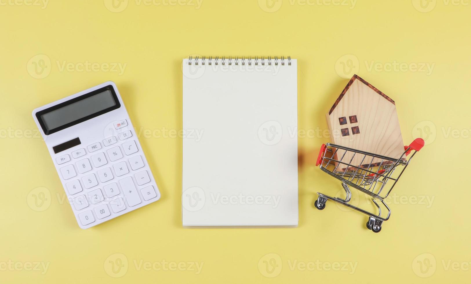 plano diseño de de madera casa modelo en compras carretilla, blanco calculadora, blanco página abrió cuaderno en amarillo antecedentes con Copiar espacio, hogar compra concepto. foto