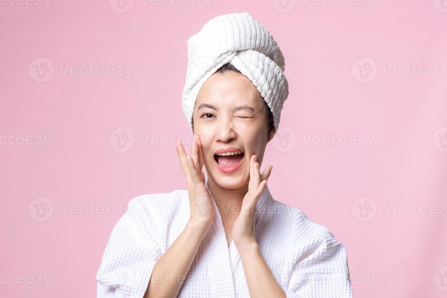 hermosa joven asiático mujer contento con limpiar cara piel en toalla y bata de baño, spa traje en rosado antecedentes. protección de la piel, tratamiento, bienestar terapia, facial cuidado, belleza hembra salud, cosmetología concepto foto