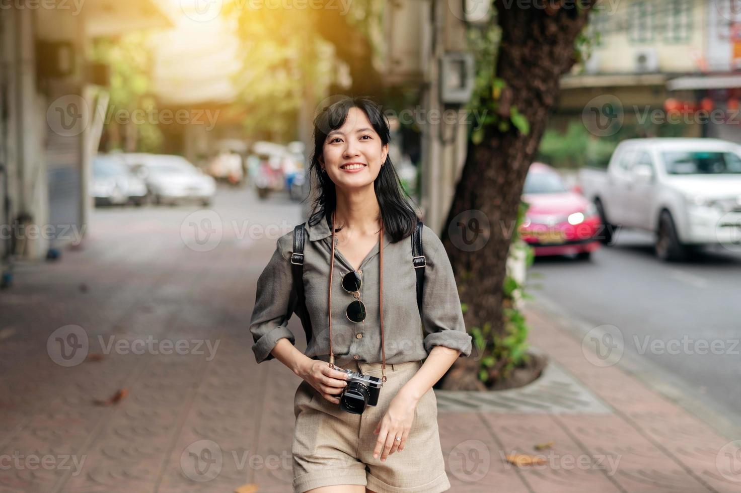 joven asiático mujer mochila viajero utilizando digital compacto cámara, disfrutando calle cultural local sitio y sonrisa. viajero comprobación fuera lado calles foto