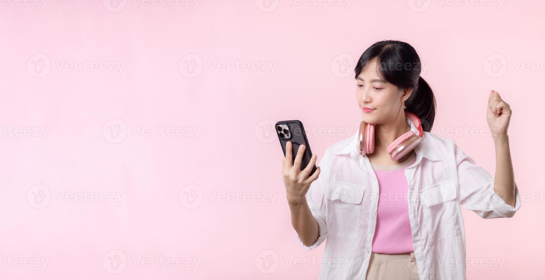 Portrait young attractive asian woman happy smile using smartphone with earphone, headphone isolated on pink studio background. Pretty female person using mobile phone. Music online lifestyle concept. photo