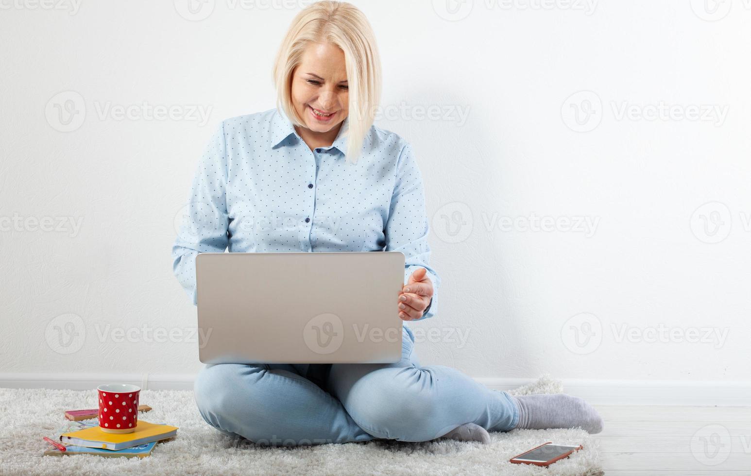 Happy middle aged woman sitting relaxed on the floor using laptop for entertainment. The concept of leisure and work with a cup of coffee at home. photo