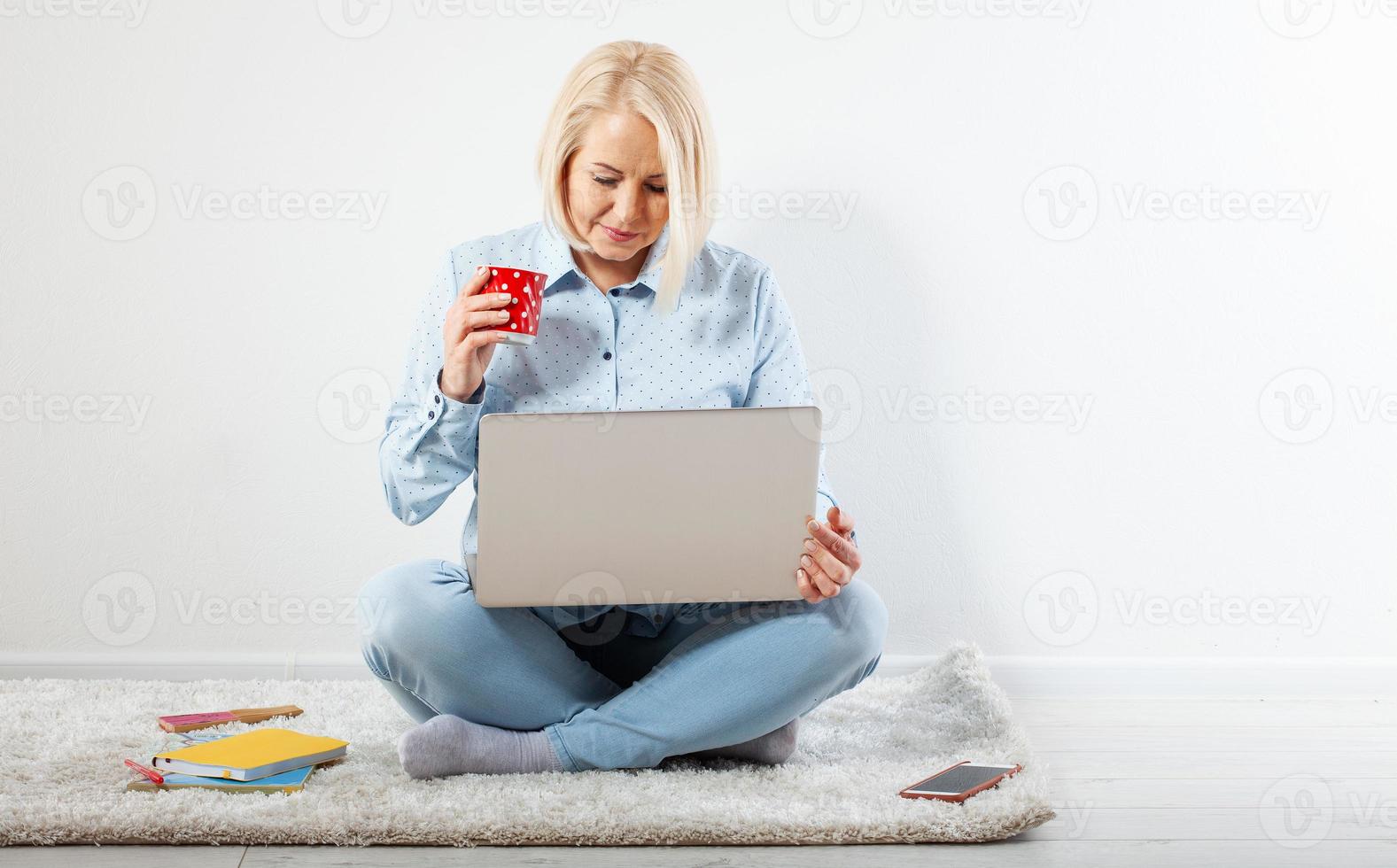 Happy middle aged woman sitting relaxed on the floor using laptop for entertainment. The concept of leisure and work with a cup of coffee at home. photo