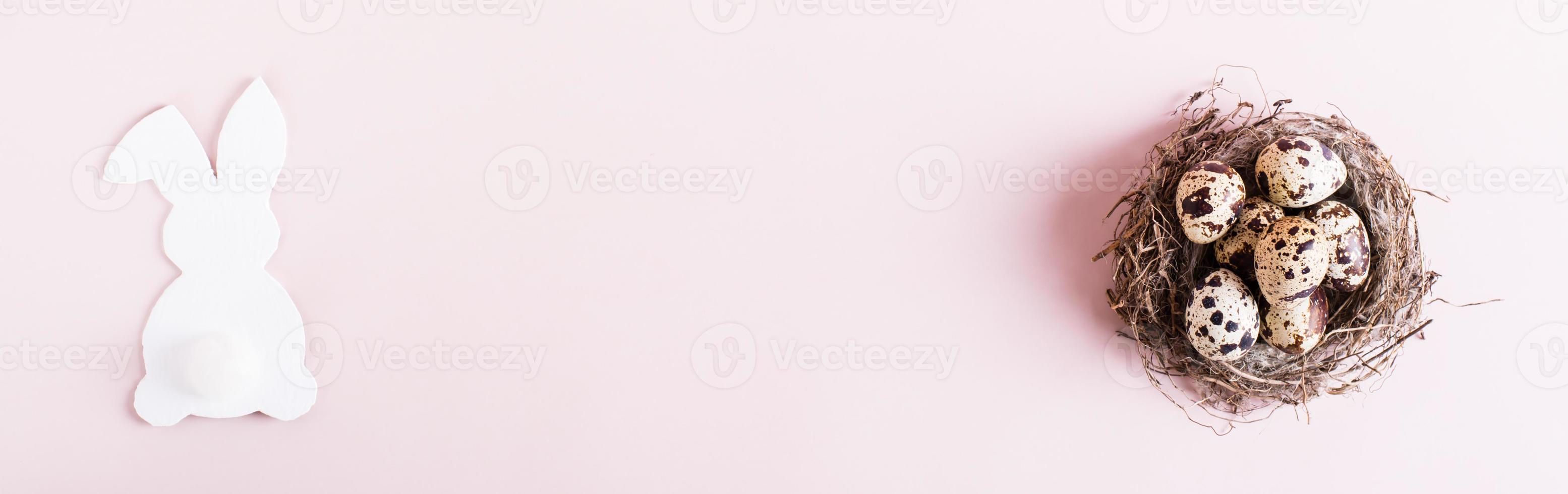 Web banner happy easter quail eggs in natural nest and white rabbit on pink background top view photo