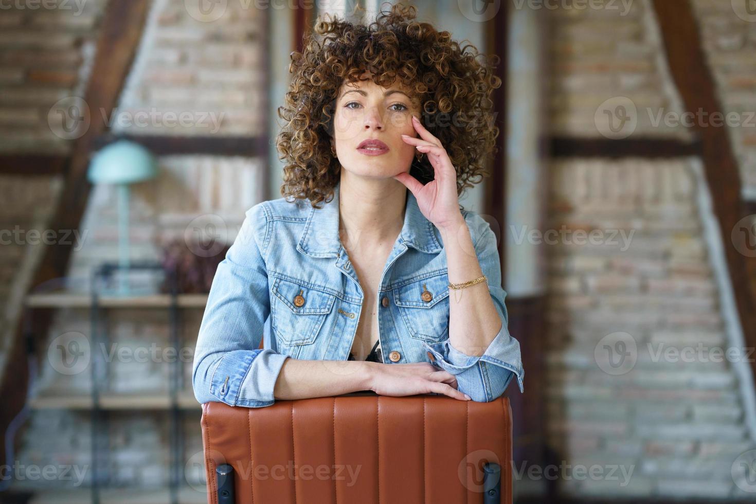 Sensual curly haired woman sitting on chair against brick wall photo