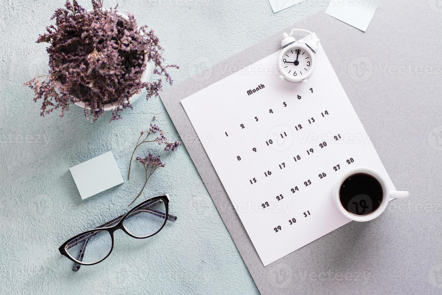 mensual calendario hoja, café taza, alarma reloj, lentes y un ramo de flores de flores en el mesa. hora planificación y organización foto