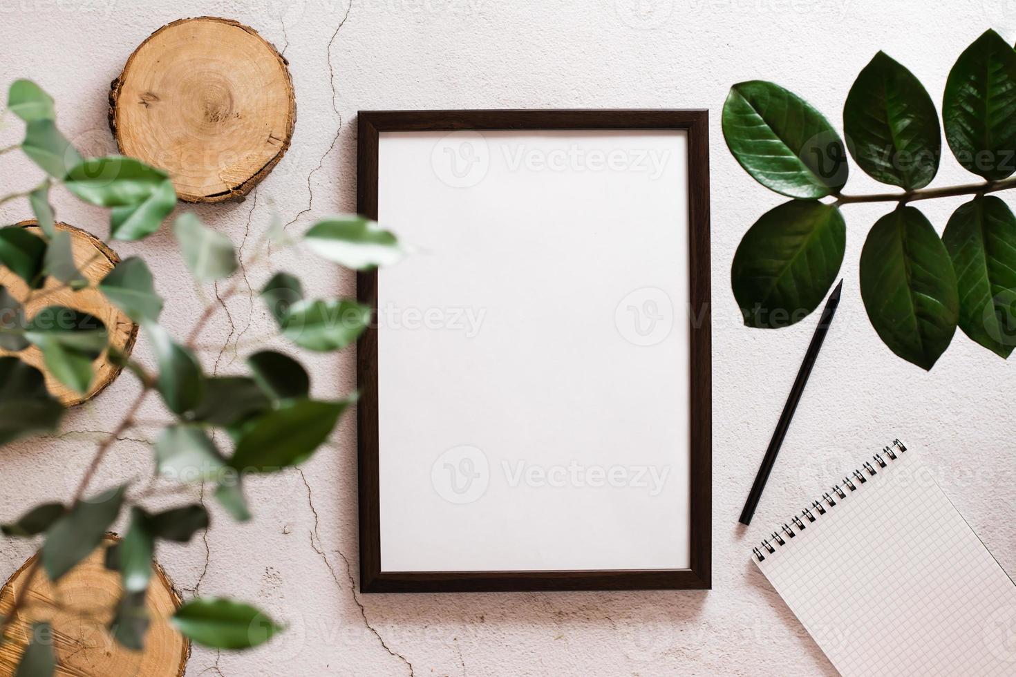 Eco desk. Blank frame with white sheet, notepad, pencil and cuts of tree trunk on light concrete background and home plant leaves. Top view. photo