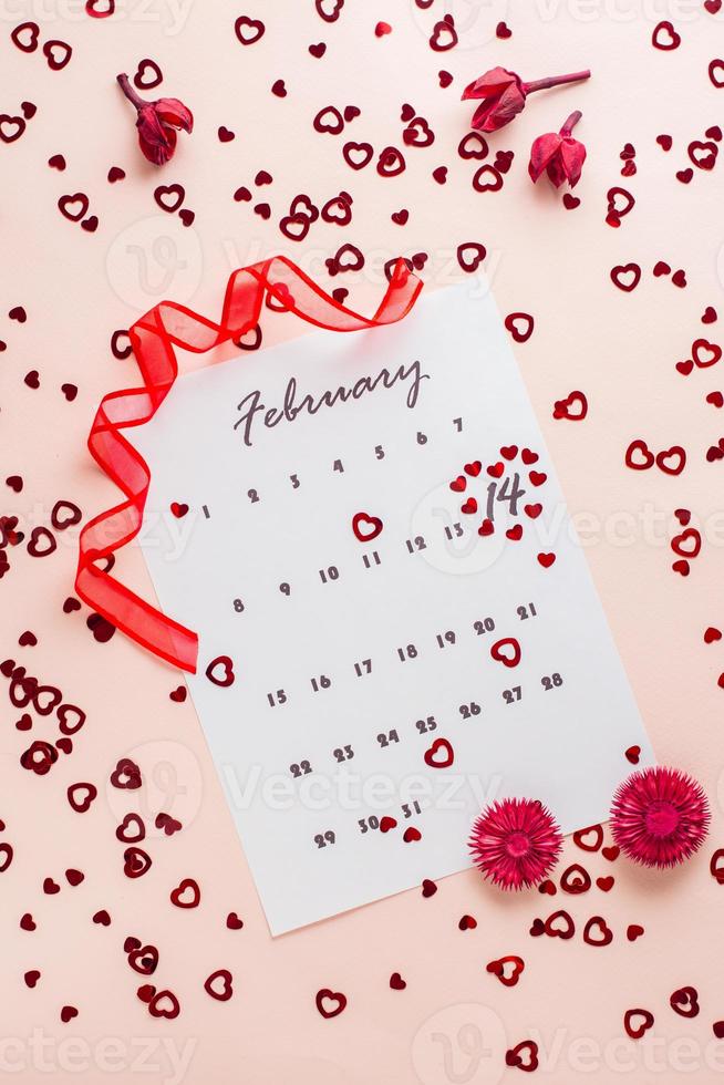 Valentine's Day. Small red hearts highlight the date February 14 on a calendar sheet, dry flowers and a red ribbon on a pink heart-strewn background. Vertical view photo
