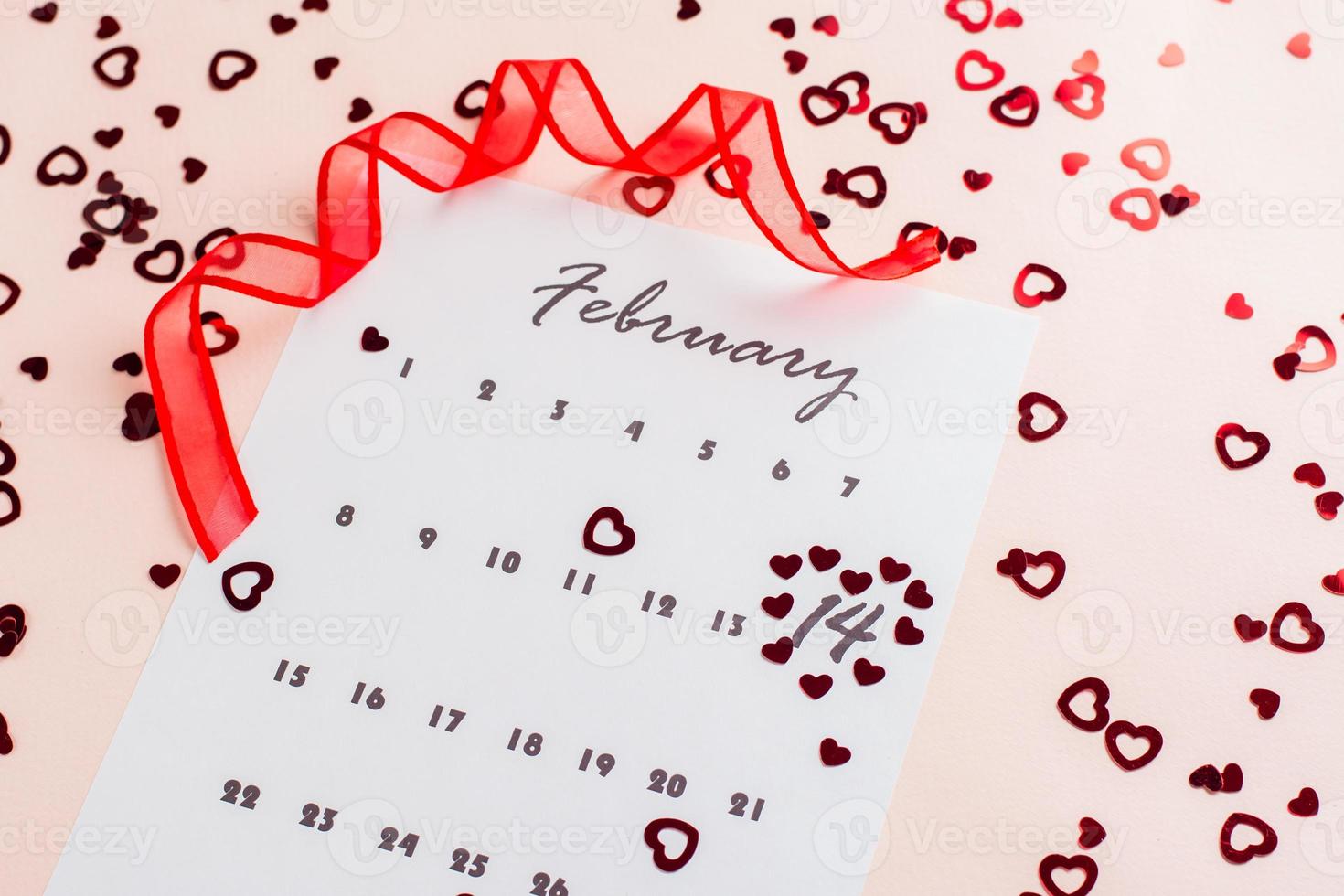 Valentine's Day. Small red hearts highlight the date February 14 on a calendar sheet and a red ribbon on a pink heart-strewn background. Close-up photo