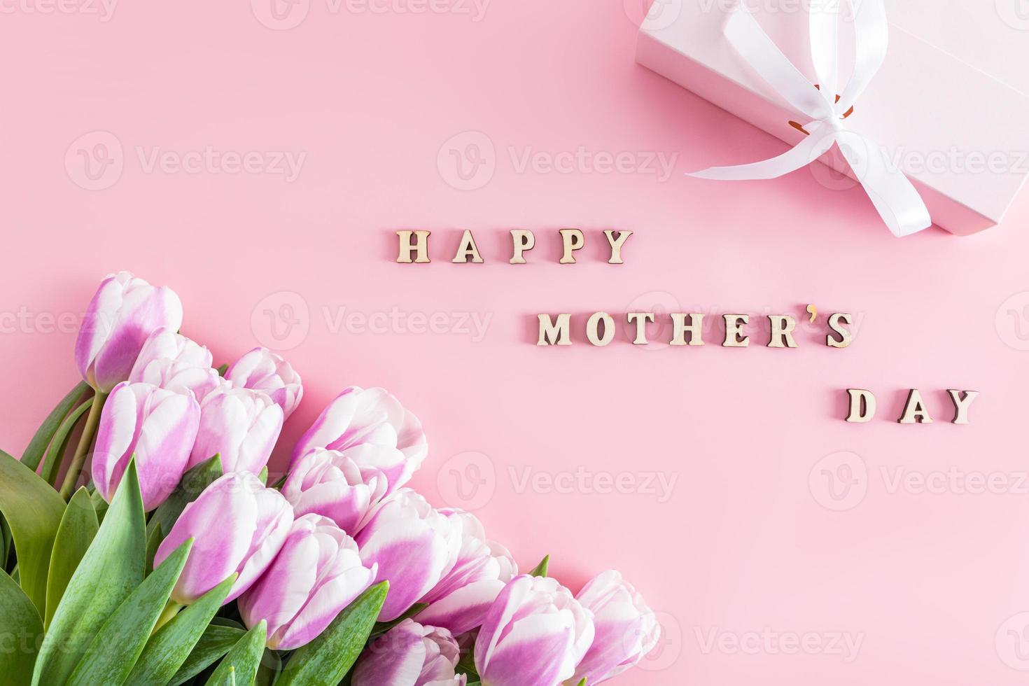 top view of a bouquet of spring tulip flowers and a festive box. text in wooden letters of Happy Mother's Day. pink background. postcard. photo