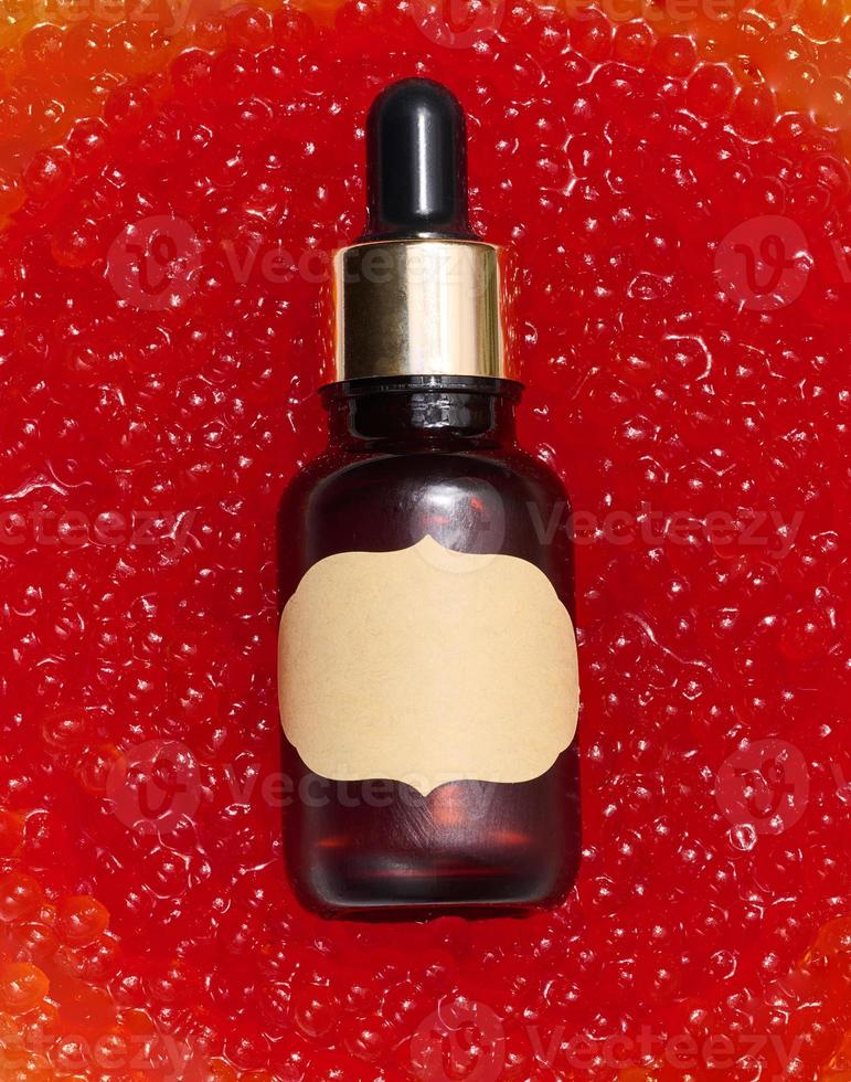 A glass brown bottle with a paper label and a pipette lies in red caviar, top view photo