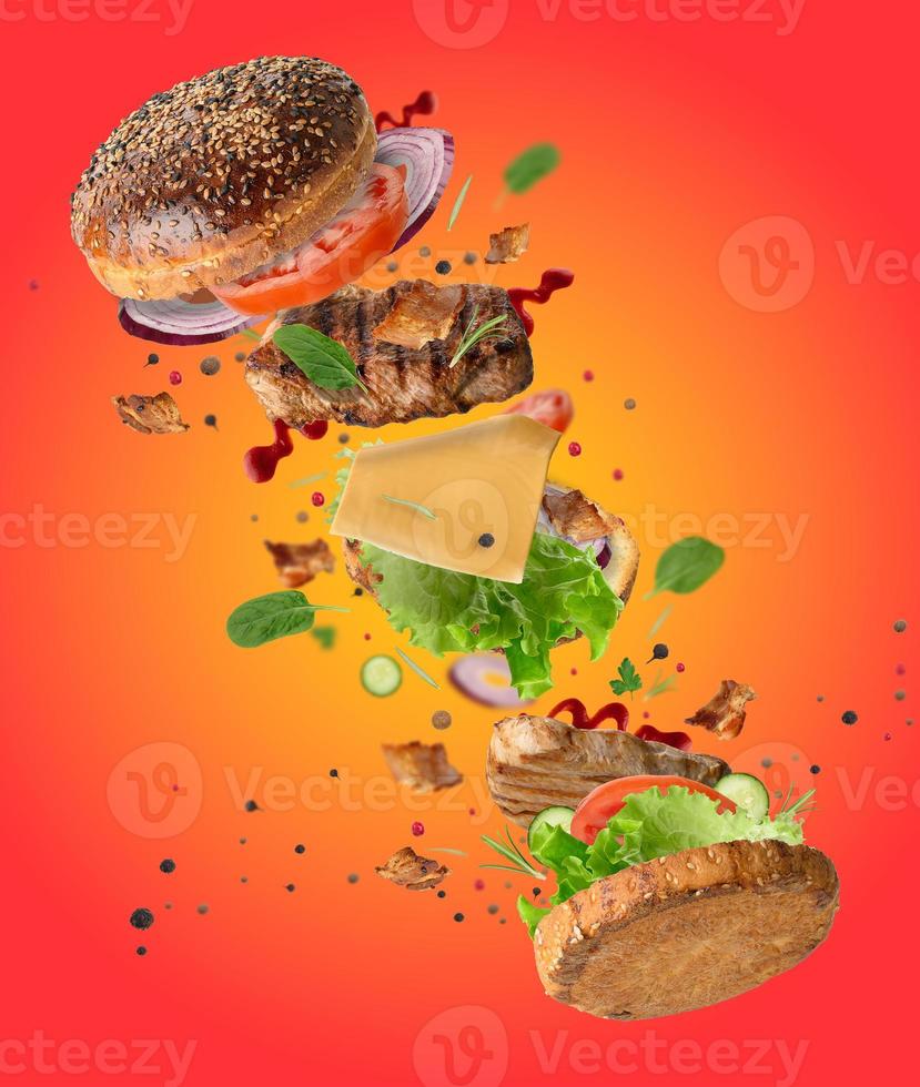 Delicious burger with flying ingredients, pieces of vegetables, spices and meat fried steak on orange background photo