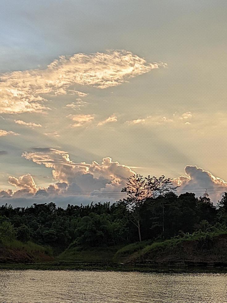 A boat is in the water and the sky is blue and the water is calm. A tree in the foreground with the sun shining through the clouds. The water is calm. photo