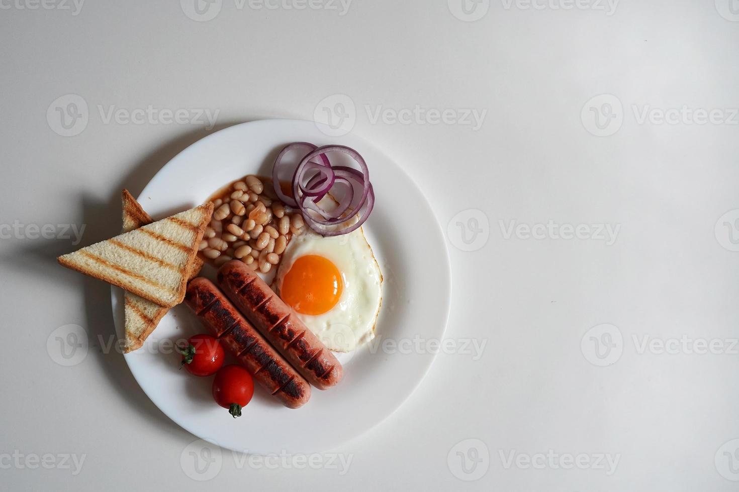 parte superior ver Inglés desayuno. frito huevo, dos frito salchichas, A la parrilla un pan tostada, Enlatado frijoles, azul cebollas y Cereza Tomates en un blanco antecedentes foto