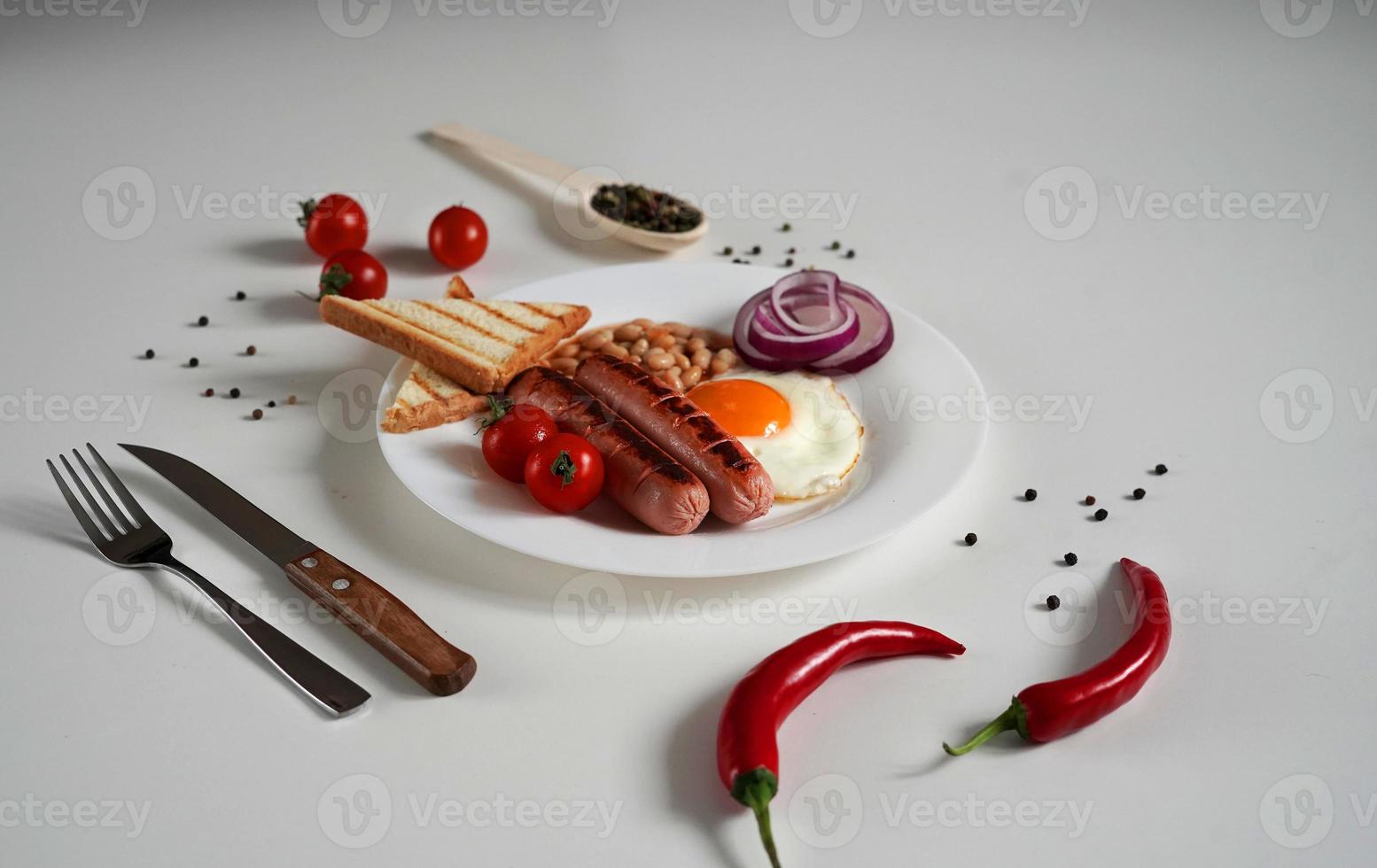 Traditional English Breakfast. Plate with Fried egg, two fried sausages, grilled bread toast, canned beans, blue onions and cherry tomatoes on a White Background. Copy space photo