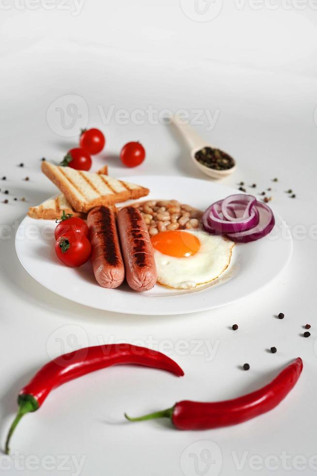 Traditional English Breakfast. Plate with Fried egg, two fried sausages, grilled bread toast, canned beans, blue onions and cherry tomatoes on a White Background. Copy space photo