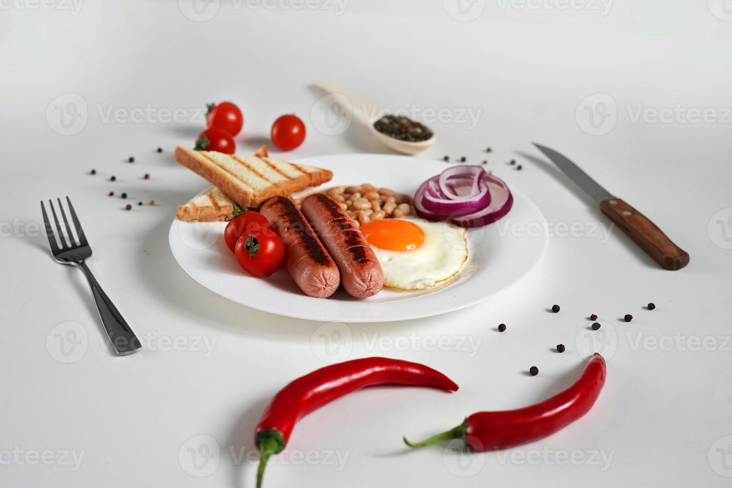 Traditional English Breakfast. Plate with Fried egg, two fried sausages, grilled bread toast, canned beans, blue onions and cherry tomatoes on a White Background. Copy space photo