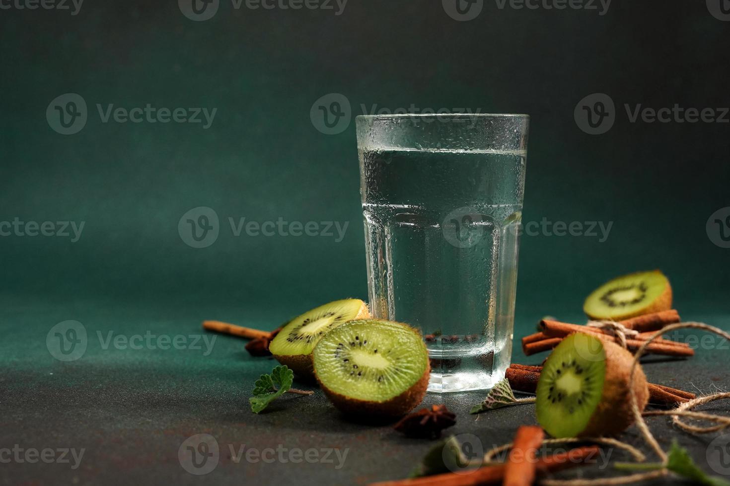 un vaso de frío agua en un verde antecedentes. decorado con rebanada kiwi, canela palos, badyan, menta hojas. Copiar espacio foto
