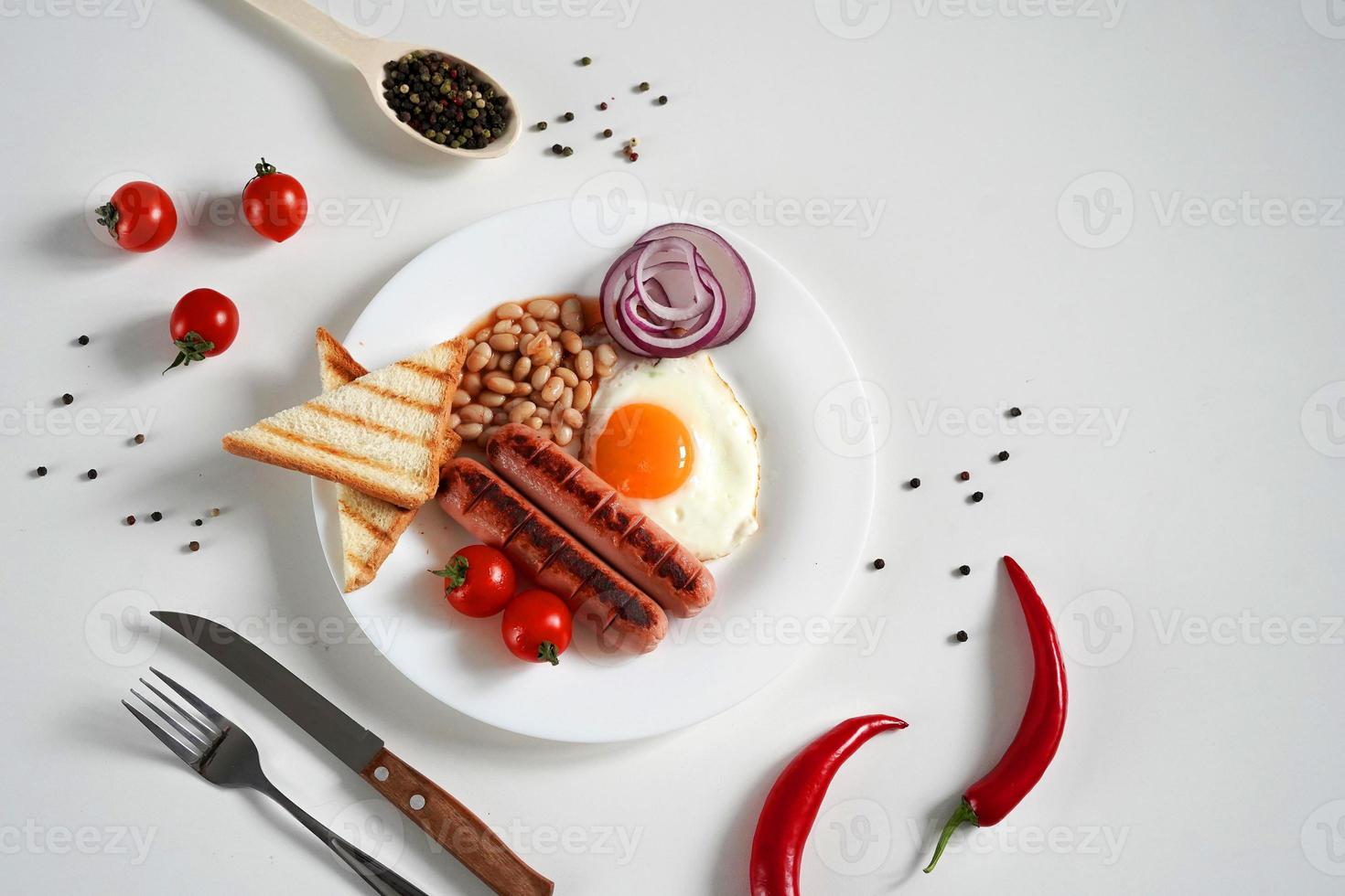 Traditional English Breakfast. Plate with Fried egg, two fried sausages, grilled bread toast, canned beans, blue onions and cherry tomatoes on a White Background. Copy space photo