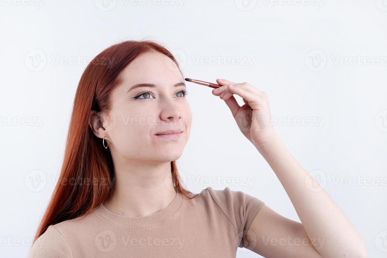 Joyful redhead young woman using makeup brush making up isolated on white background photo