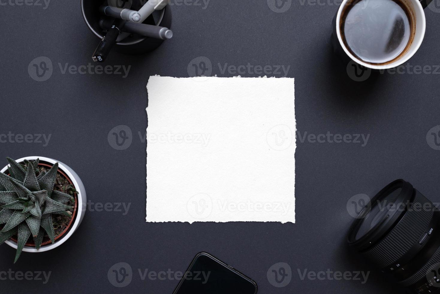 Blank paper frame on black office desk surrounded by phone, camera, coffee mug, plant and pens. Top view, flat lay composition for copy presentation photo