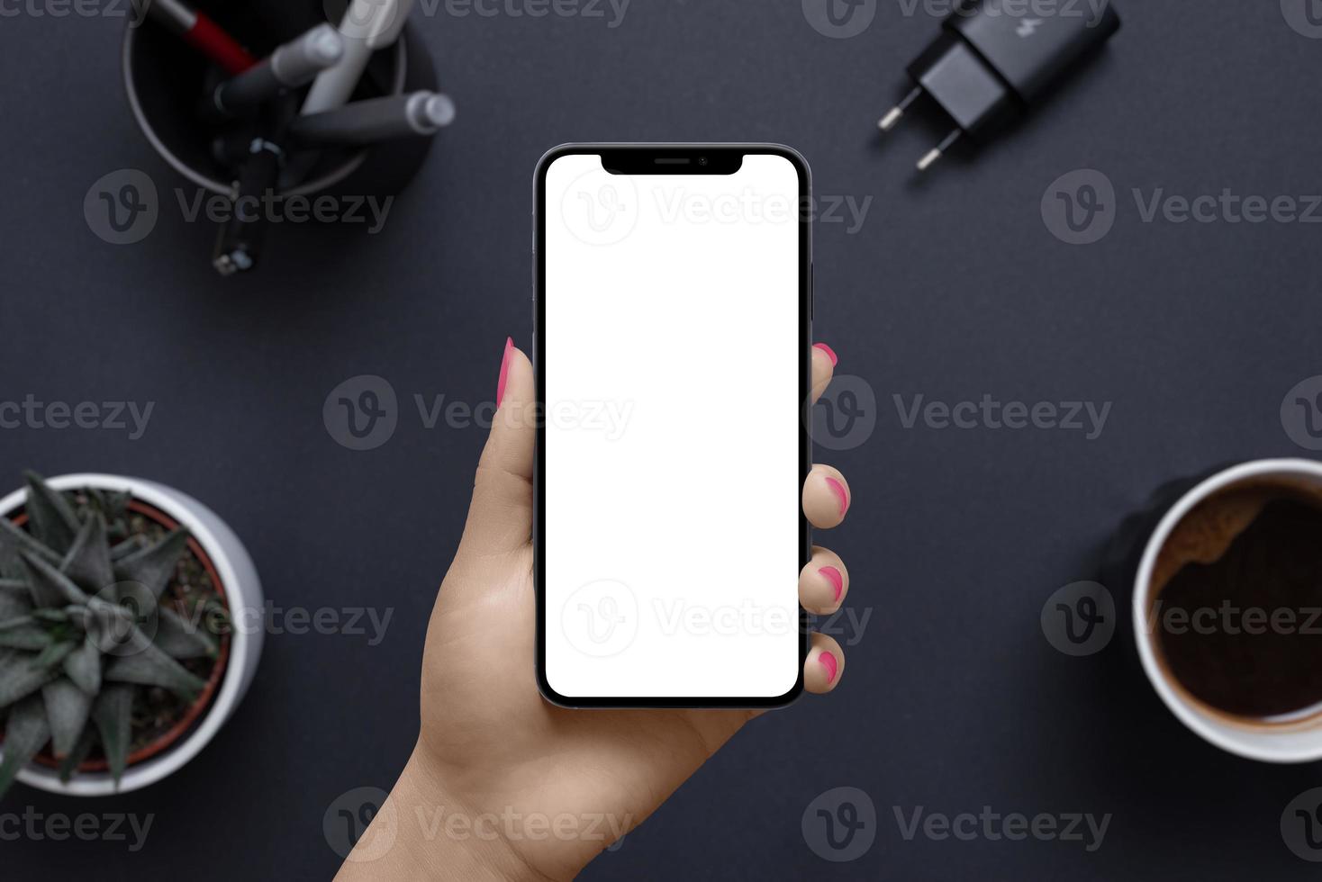 Phone in woman hand with isolated display for mockup. Business, office mockup for app or web page presentation. Office desk with coffee mug, plant, pens, charger in background photo