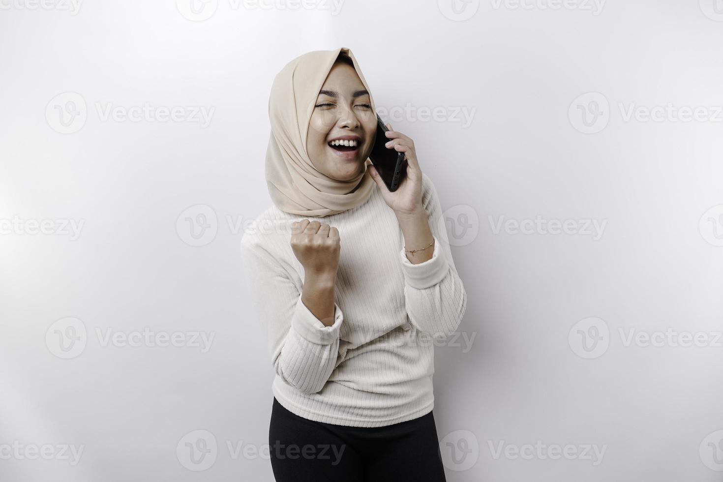un contento asiático musulmán mujer vistiendo un Pañuelo, participación su teléfono, aislado por blanco antecedentes foto