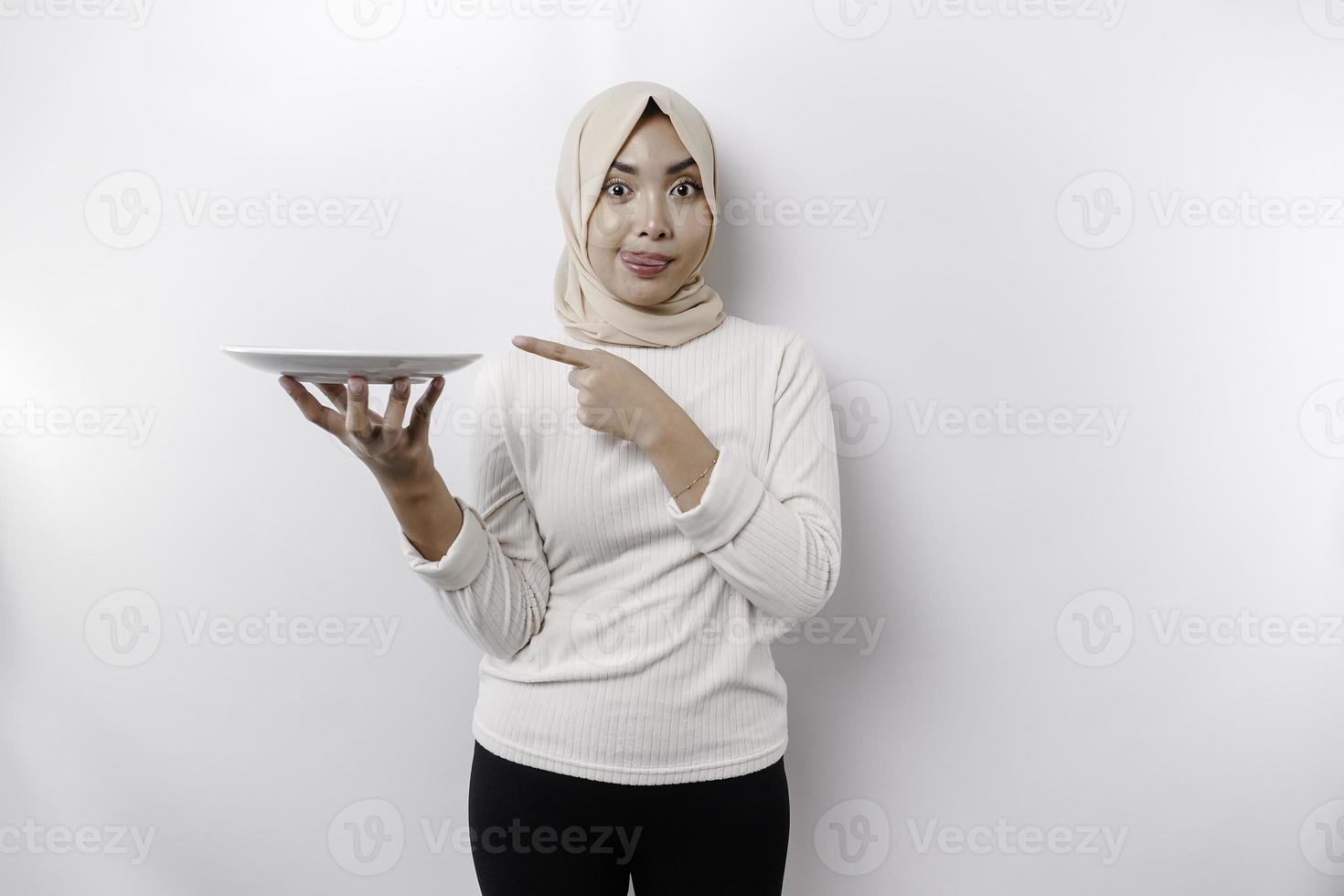 A smiling Asian Muslim woman is fasting and hungry and holding and pointing to a plate photo