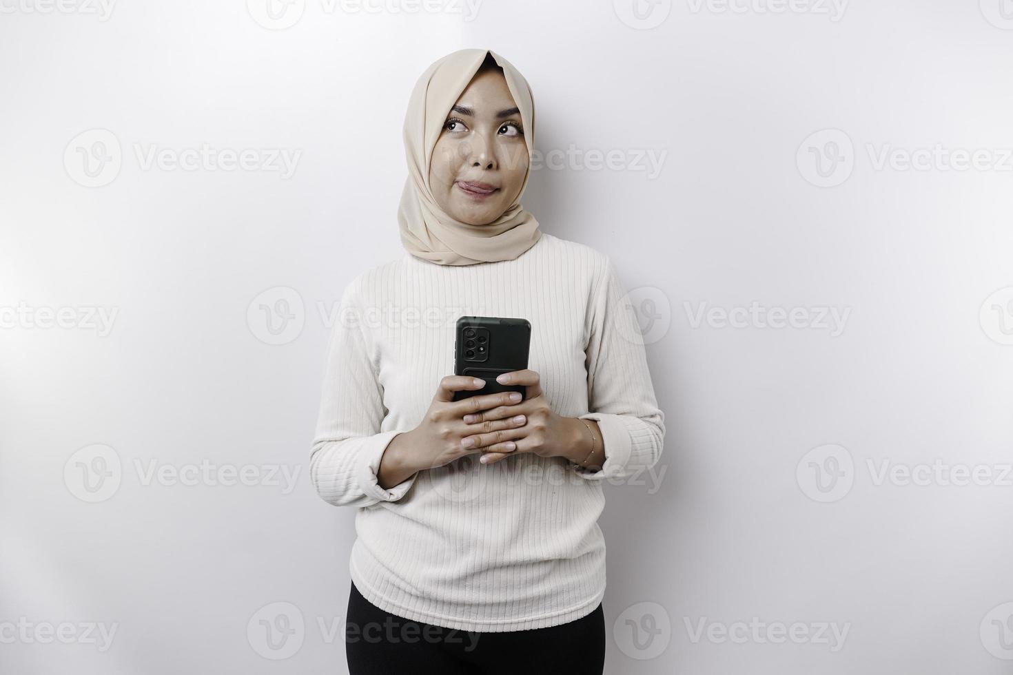 un pensativo joven asiático musulmán mujer vistiendo un hijab y participación su barbilla mientras hablando en el teléfono aislado por un blanco antecedentes foto