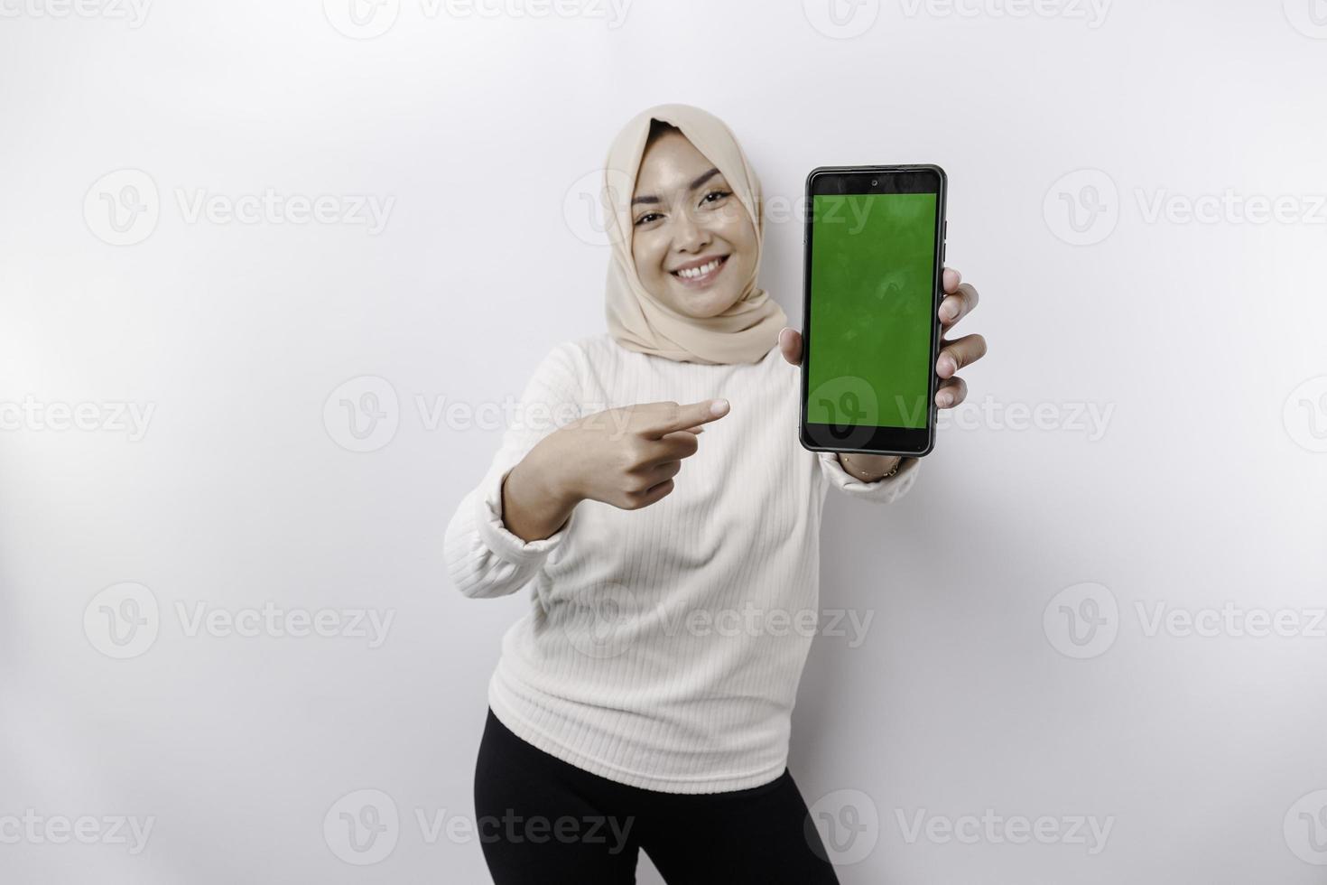 A happy Asian Muslim woman wearing a headscarf, showing her phone screen, isolated by white background photo