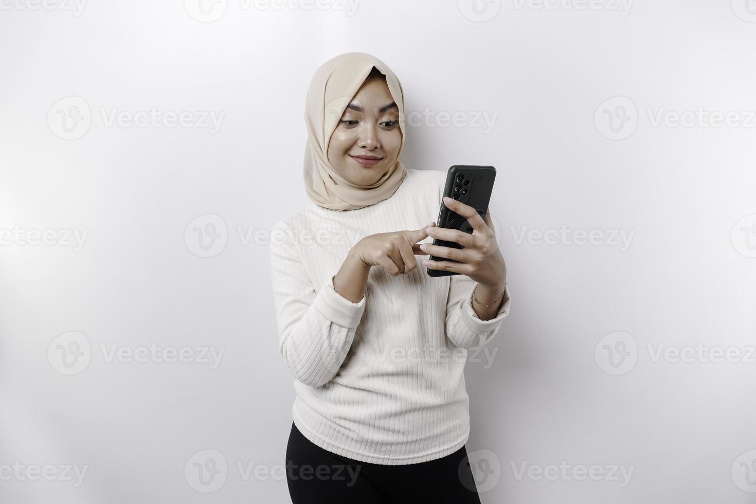 un contento asiático musulmán mujer vistiendo un Pañuelo, participación su teléfono, aislado por blanco antecedentes foto