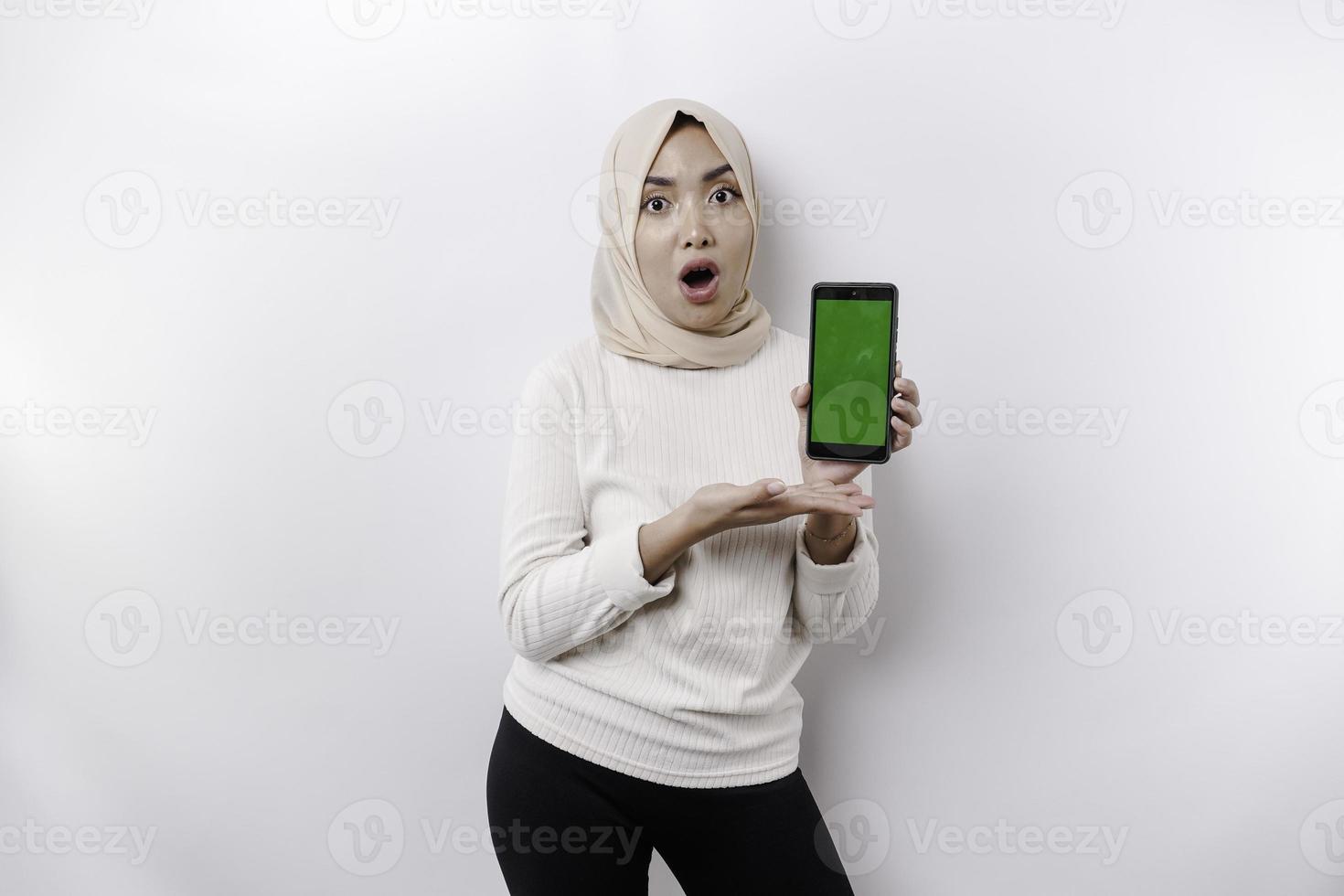Shocked Asian woman wearing headscarf, showing copy space on her phone screen, isolated by white background photo