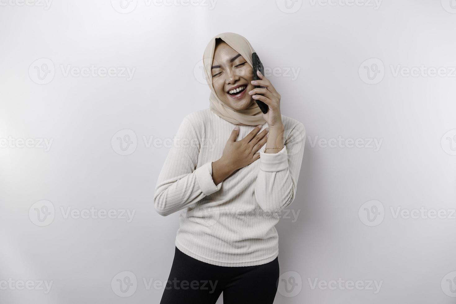 A happy Asian Muslim woman wearing a headscarf, holding her phone, isolated by white background photo