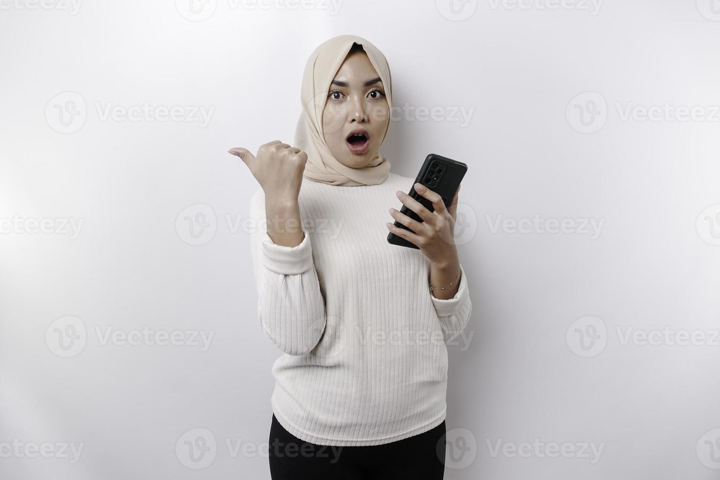 Shocked Asian Muslim woman wearing hijab pointing at the copy space beside her while holding her phone, isolated by white background photo