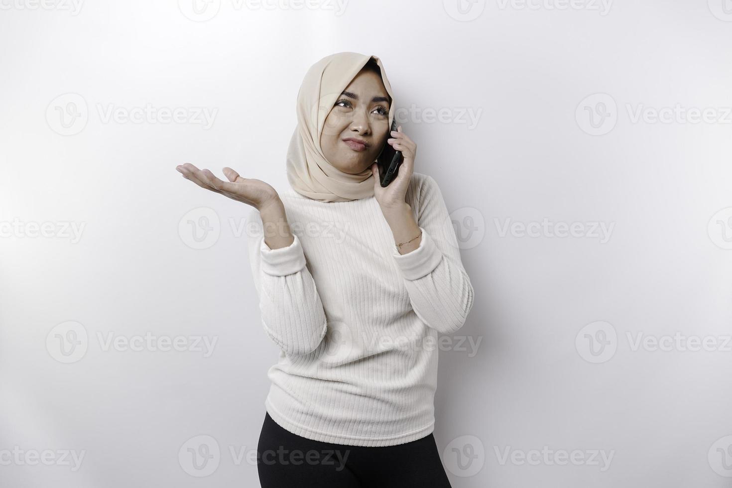 A thoughtful young Asian Muslim woman wearing a hijab and pointing copy space beside her while talking on the phone isolated by a white background photo