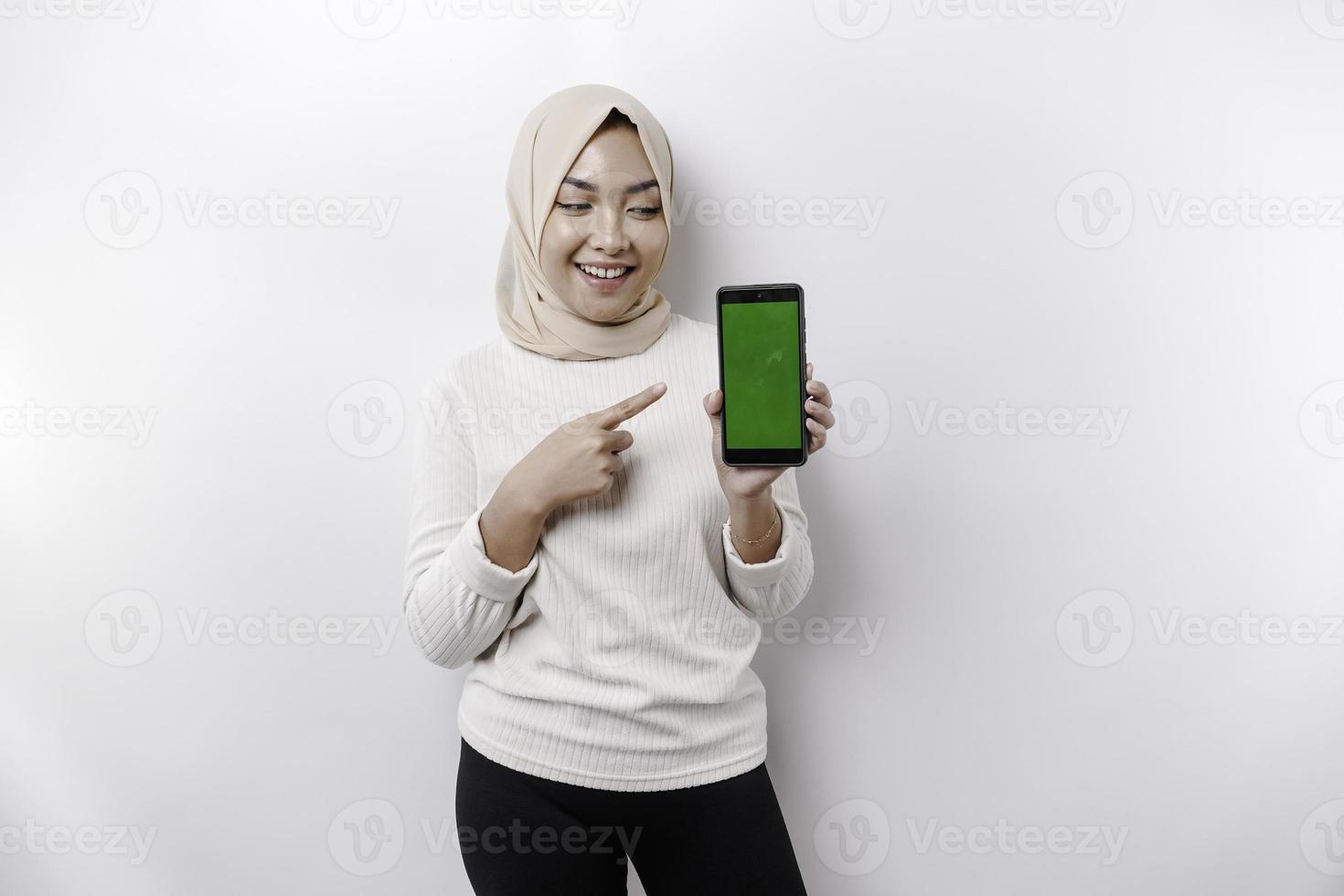 A happy Asian Muslim woman wearing a headscarf, showing her phone screen, isolated by white background photo