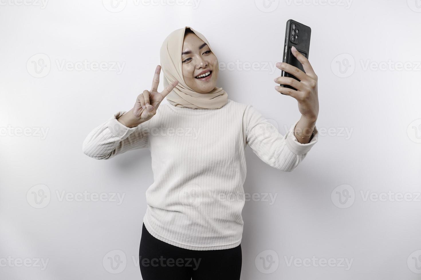 un contento asiático musulmán mujer vistiendo un Pañuelo, participación su teléfono, aislado por blanco antecedentes foto