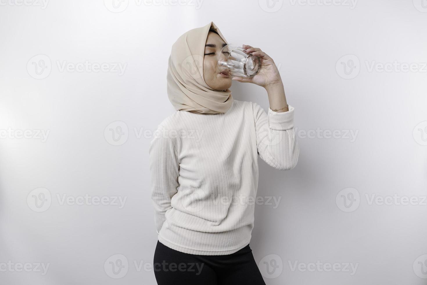 Joyful Asian Muslim woman wearing headscarf is drinking a glass of water, isolated on white background. photo