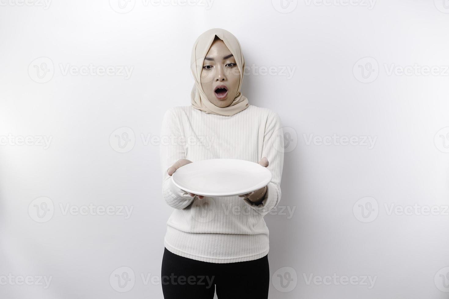 A shocked Asian Muslim woman is fasting and hungry and holding a plate with her mouth wide open photo