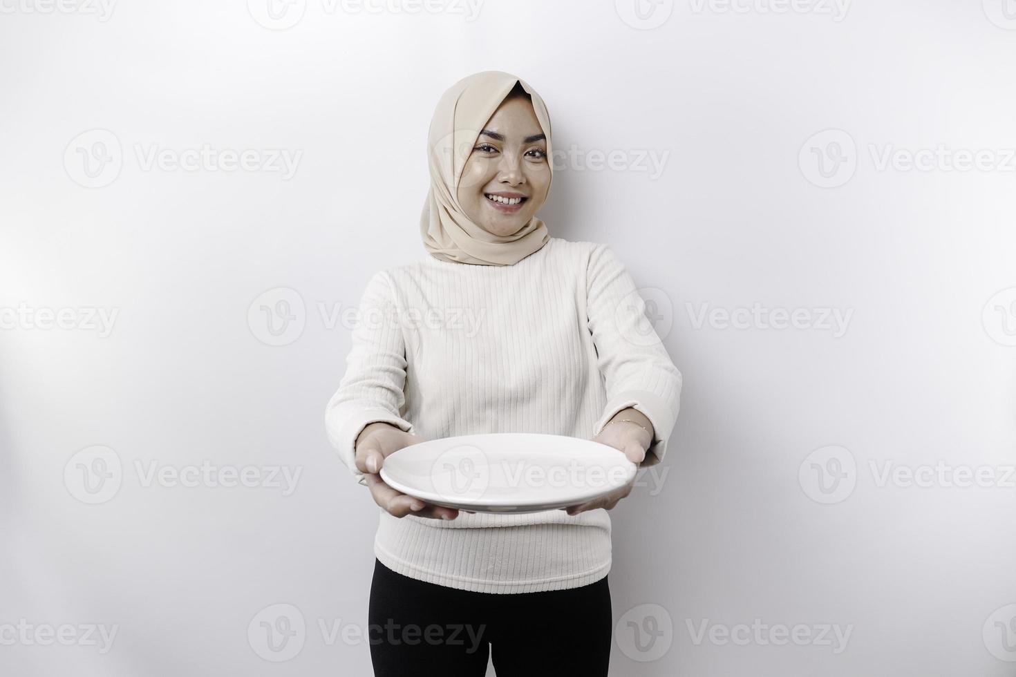 A smiling Asian Muslim woman is fasting and hungry and holding and pointing to a plate photo